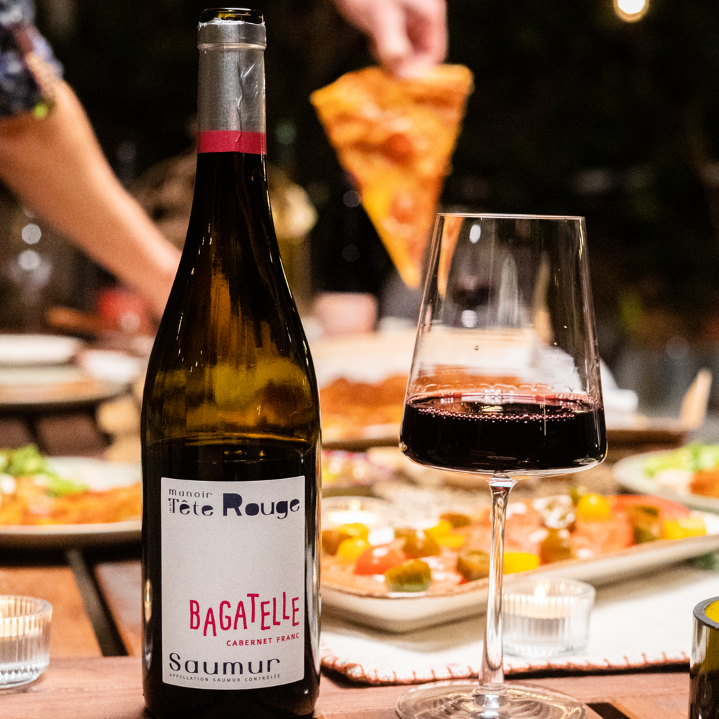 A bottle and glass of wine next to each other filled with red wine, with food in the background.