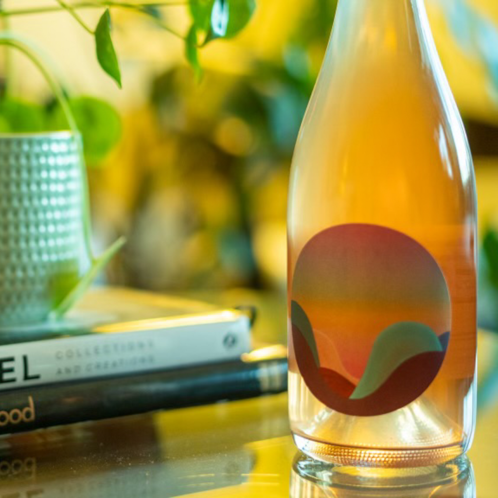 A picture of an orange wine bottle on a table next to books and a plant.