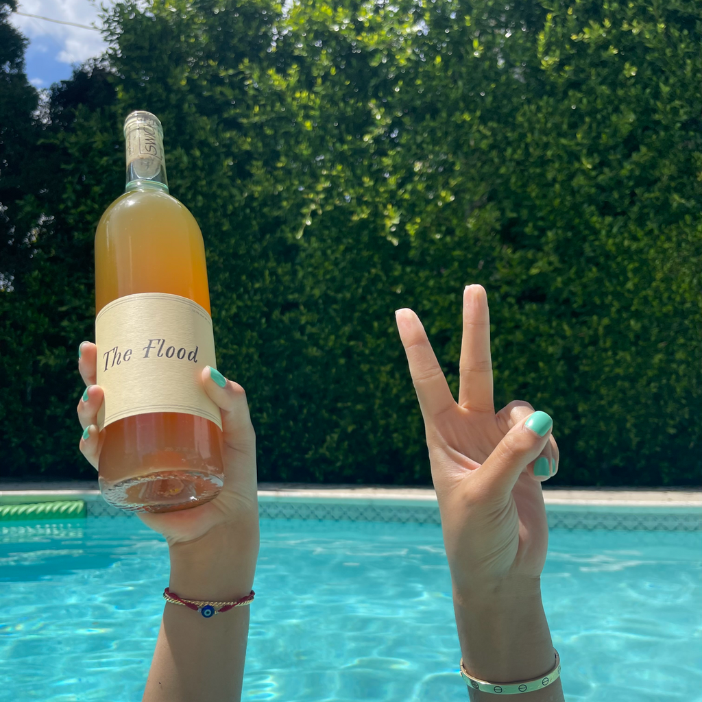 A picture shot in the pool, of a woman holding a bottle of orange wine in one hand and showing the peace sign with the other hand. 