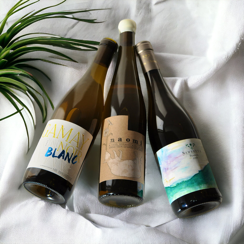 Three bottles of white wine laying down together on a white silk cloth, with a plant next to them.