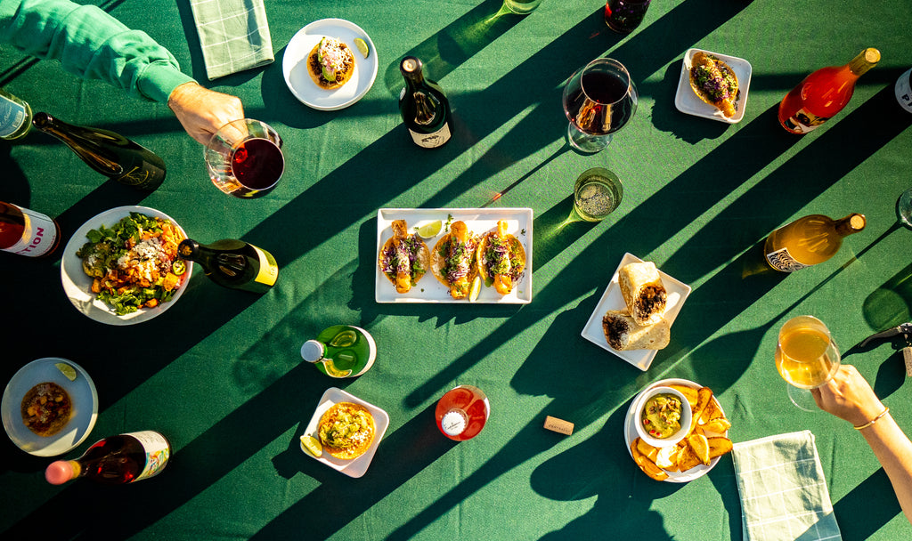 Table with green table cloth displaying mexican food and natural wines as well as two people holding wine glasses toasting one another