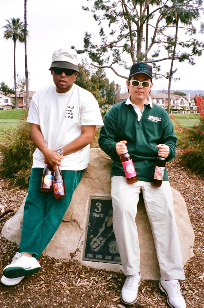 A photo of two men dressed in a hip golf attire (bucket hat, trucker hat, sunglasses, polo, t-shirt, golf pants and sneakers) in white and green colors, each holding two bottles of red wine, while sitting on a rock monument with the golf course and palm trees in the background.