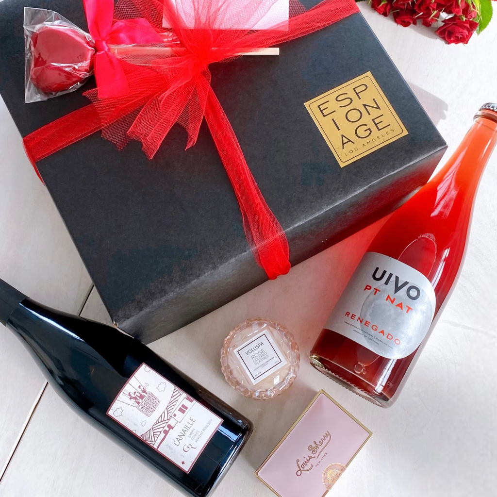 A photo of the Unfined Wines Valentine's Day GIft Box which shows a black box wrapped in red bow that comes with a red heart lollipop, a personalized white card, two bottles of red wine, a soap, chocolates and roses.